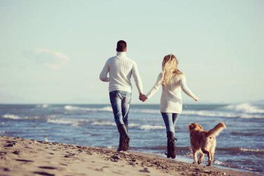 Plajda koşan çift, sonbahar gününde köpekle el ele tutuşuyor.