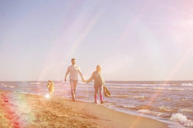 Plajda koşan çift, sonbahar gününde köpekle el ele tutuşuyor.