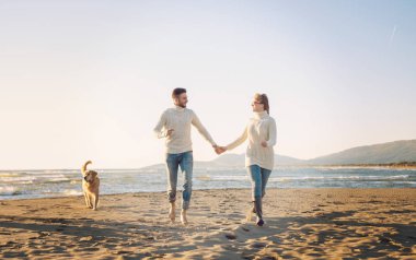 Plajda koşan çift, sonbahar gününde köpekle el ele tutuşuyor.