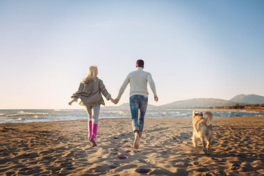 Couple Running On The Beach Holding Their Hands with dog On autmun day clipart