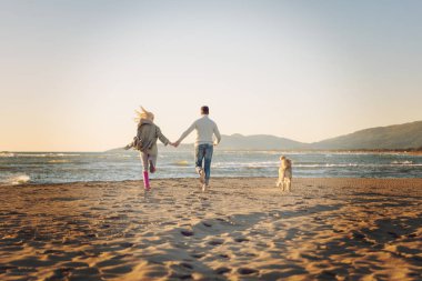 Plajda koşan çift, sonbahar gününde köpekle el ele tutuşuyor.