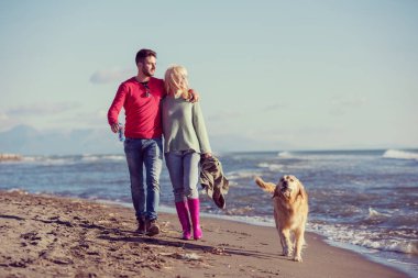 Plajda koşan çift, sonbahar gününde köpekle el ele tutuşuyor.