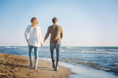 Genç çift, sonbahar güneşli bir günde kumsalda yürüyüp sarılıyor.