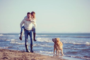 Plajda koşan çift, sonbahar gününde köpekle el ele tutuşuyor.