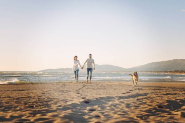 Plajda koşan çift, sonbahar gününde köpekle el ele tutuşuyor.