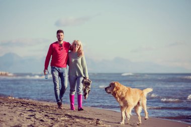Plajda koşan çift, sonbahar gününde köpekle el ele tutuşuyor.