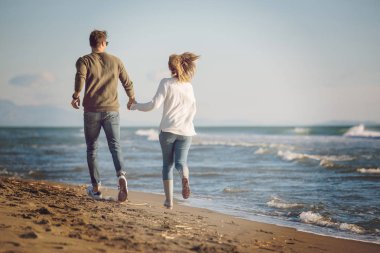 Genç çift, sonbahar güneşli bir günde kumsalda yürüyüp sarılıyor.