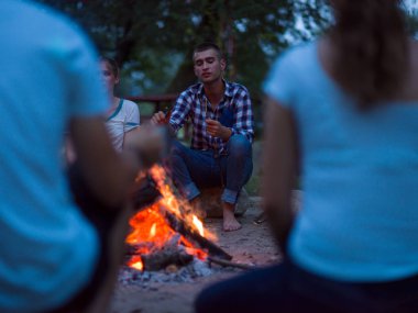 Bir grup mutlu genç arkadaş nehir kıyısında kamp ateşi etrafında dinleniyor ve eğleniyorlar.