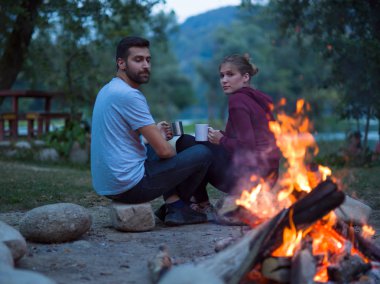 Bir bardak çay veya kahve kendilerini ısınma tutan akşam kamp ateşi etrafında oturan genç çift