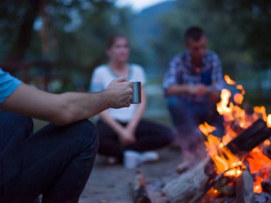Bir grup mutlu genç arkadaş nehir kıyısında kamp ateşi etrafında dinleniyor ve eğleniyorlar.