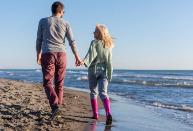 Genç çift, sonbahar güneşli bir günde kumsalda yürüyüp sarılıyor.