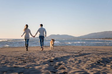 Plajda koşan çift, sonbahar gününde köpekle el ele tutuşuyor.