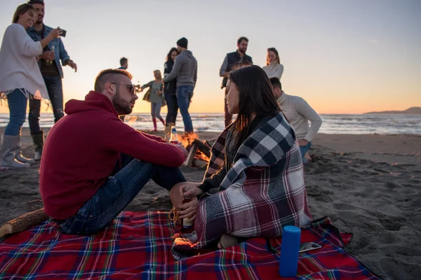 ビーチでキャンプファイヤーの周りの友人と楽しむ若いカップル日没時にビールを飲む — ストック写真