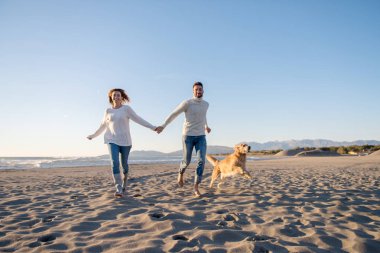 Plajda koşan çift, sonbahar gününde köpekle el ele tutuşuyor.