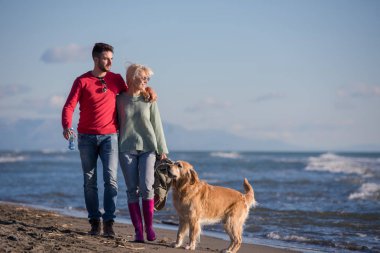 Plajda koşan çift, sonbahar gününde köpekle el ele tutuşuyor.