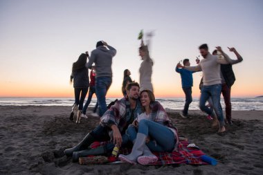 Genç çift gün batımında arkadaşlarıyla kumsaldaki kamp ateşinde bira içerek eğleniyor.