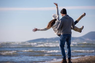 Genç çift, sonbahar güneşli bir günde kumsalda yürüyüp sarılıyor.