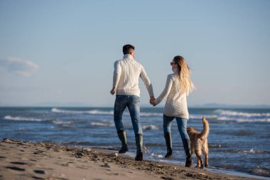 Plajda koşan çift, sonbahar gününde köpekle el ele tutuşuyor.