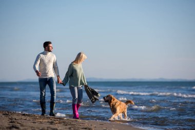 Plajda koşan çift, sonbahar gününde köpekle el ele tutuşuyor.