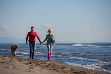 Plajda koşan çift, sonbahar gününde köpekle el ele tutuşuyor.