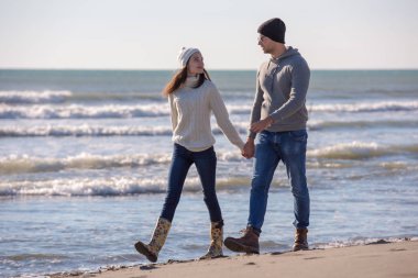 Genç çift, sonbahar güneşli bir günde kumsalda yürüyüp sarılıyor.