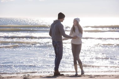 Genç çift, sonbahar güneşli bir günde kumsalda yürüyüp sarılıyor.