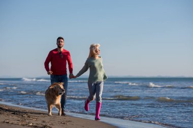 Plajda koşan çift, sonbahar gününde köpekle el ele tutuşuyor.