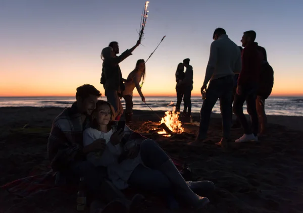 在秋天的海滩派对上 情侣们与朋友们一起喝啤酒玩乐 一起使用手机 — 图库照片