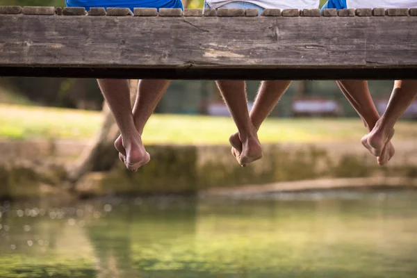 足にぶら下がっているに焦点を当てると川に架かる木製の橋で座っている人のグループ — ストック写真