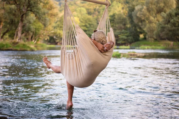 川のほとりで自然を楽しみながらハンモックで休んで若いブロンドの女性 — ストック写真