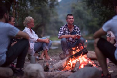 Bir grup mutlu genç arkadaş nehir kıyısında kamp ateşi etrafında dinleniyor ve eğleniyorlar.
