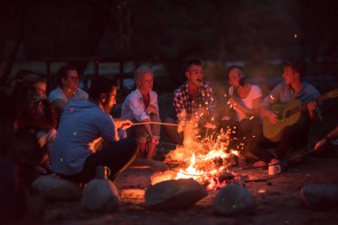 Bir grup mutlu genç arkadaş nehir kıyısında kamp ateşi etrafında dinleniyor ve eğleniyorlar.