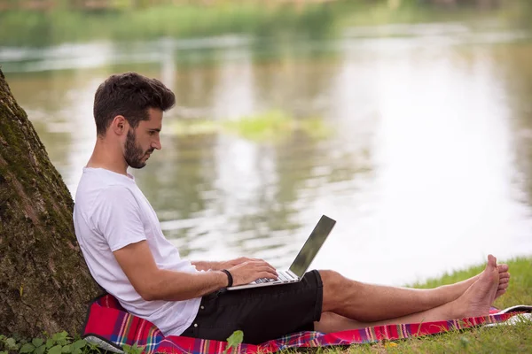 川の土手にツリーの下で美しい自然で作業しながらノート パソコンを使用して若いフリーランサー — ストック写真