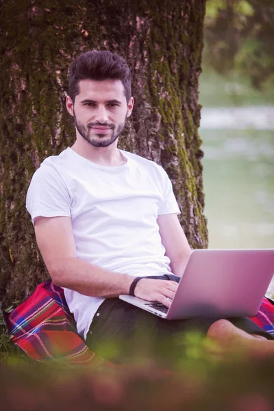 一个年轻的自由职业者使用笔记本电脑 而工作在美丽的大自然树下河岸 — 图库照片
