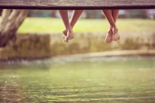 group of people sitting at wooden bridge over the river with focus on hanging legs