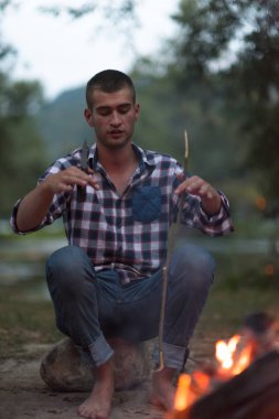 Bir grup mutlu genç arkadaş nehir kıyısında kamp ateşi etrafında dinleniyor ve eğleniyorlar.