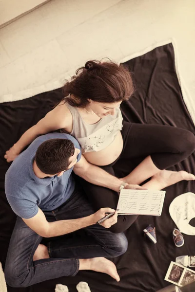 床の上に胎児の赤ちゃんを自宅のもののリストをチェック幸せな妊娠中のカップルの平面図 — ストック写真