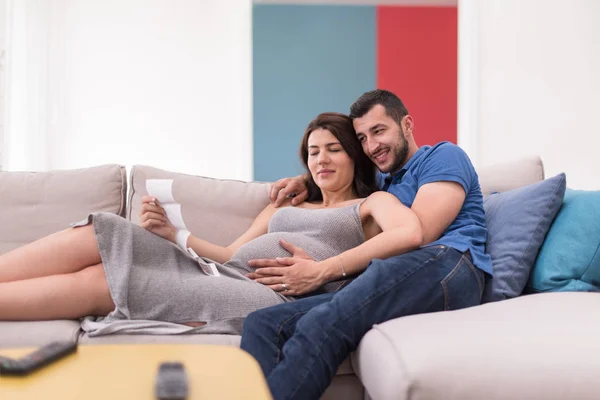自宅のソファでリラックスしながら赤ちゃんの超音波写真を見て妊娠しているカップル — ストック写真
