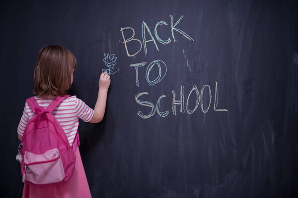 Happy school girl child with backpack writing  back to school on black chalkboard