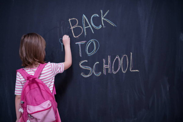 Happy school girl child with backpack writing  back to school on black chalkboard
