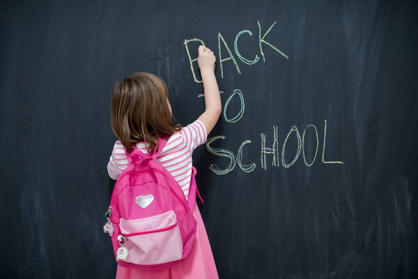 Happy school girl child with backpack writing  back to school on black chalkboard