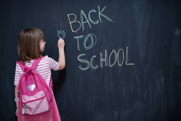 Happy school girl child with backpack writing  back to school on black chalkboard