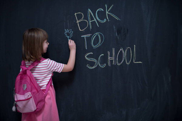 Happy school girl child with backpack writing  back to school on black chalkboard