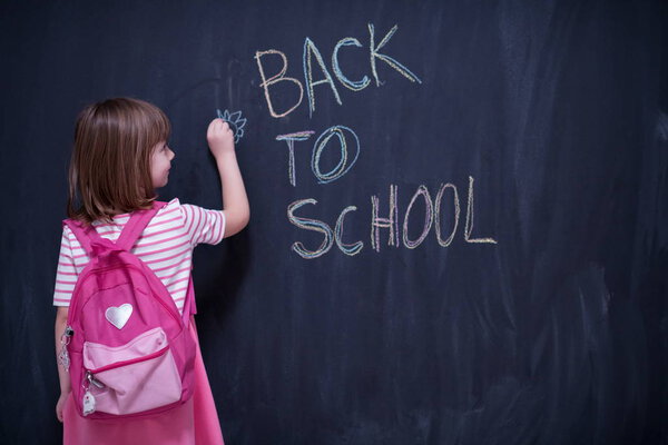 Happy school girl child with backpack writing  back to school on black chalkboard