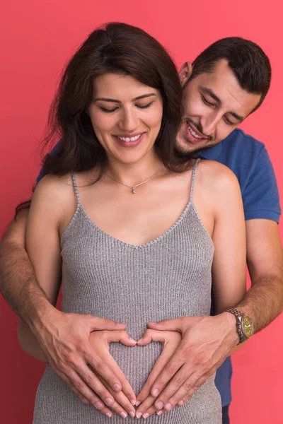 愉快的年轻夫妇显示心脏与他们的手在怀孕的妻子的肚子被隔绝在红色背景 — 图库照片