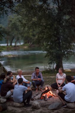 Bir grup mutlu genç arkadaş nehir kıyısında kamp ateşi etrafında dinleniyor ve eğleniyorlar.