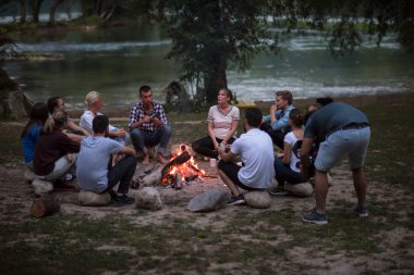 Bir grup mutlu genç arkadaş nehir kıyısında kamp ateşi etrafında dinleniyor ve eğleniyorlar.