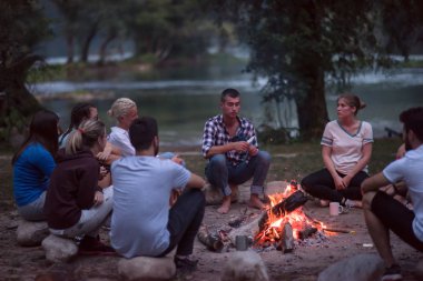Bir grup mutlu genç arkadaş nehir kıyısında kamp ateşi etrafında dinleniyor ve eğleniyorlar.
