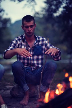 Bir grup mutlu genç arkadaş nehir kıyısında kamp ateşi etrafında dinleniyor ve eğleniyorlar.