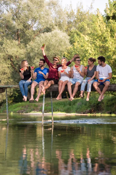 美しい自然の川に架かる木製の橋に座ってスイカを楽しんでいる若い友人のグループ — ストック写真